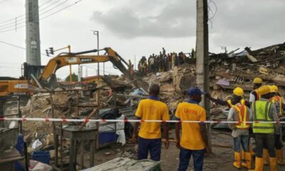 Construction workers in yellow safety vests and helmets stand among rubble as a yellow excavator clears debris at a collapsed site.
