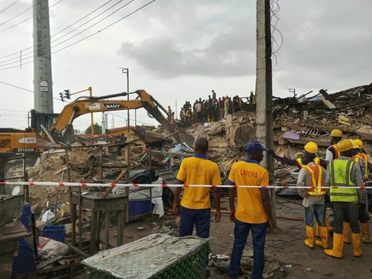Construction workers in yellow safety vests and helmets stand among rubble as a yellow excavator clears debris at a collapsed site.