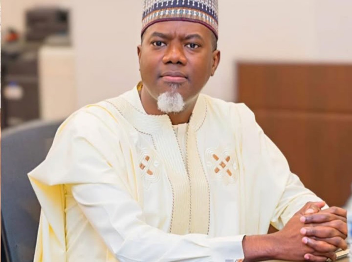 Portrait of a man wearing a white traditional outfit and patterned cap, seated at a desk with hands folded in front, in an office setting.