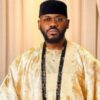Portrait of a man in a gold traditional agbada with a black cap, glasses, and long black beads.