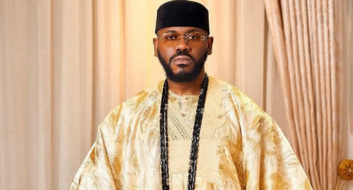Portrait of a man in a gold traditional agbada with a black cap, glasses, and long black beads.