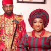 Man and woman in vibrant Nigerian wedding attire posing together; the man holds a decorative staff.