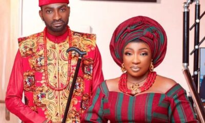 Man and woman in vibrant Nigerian wedding attire posing together; the man holds a decorative staff.