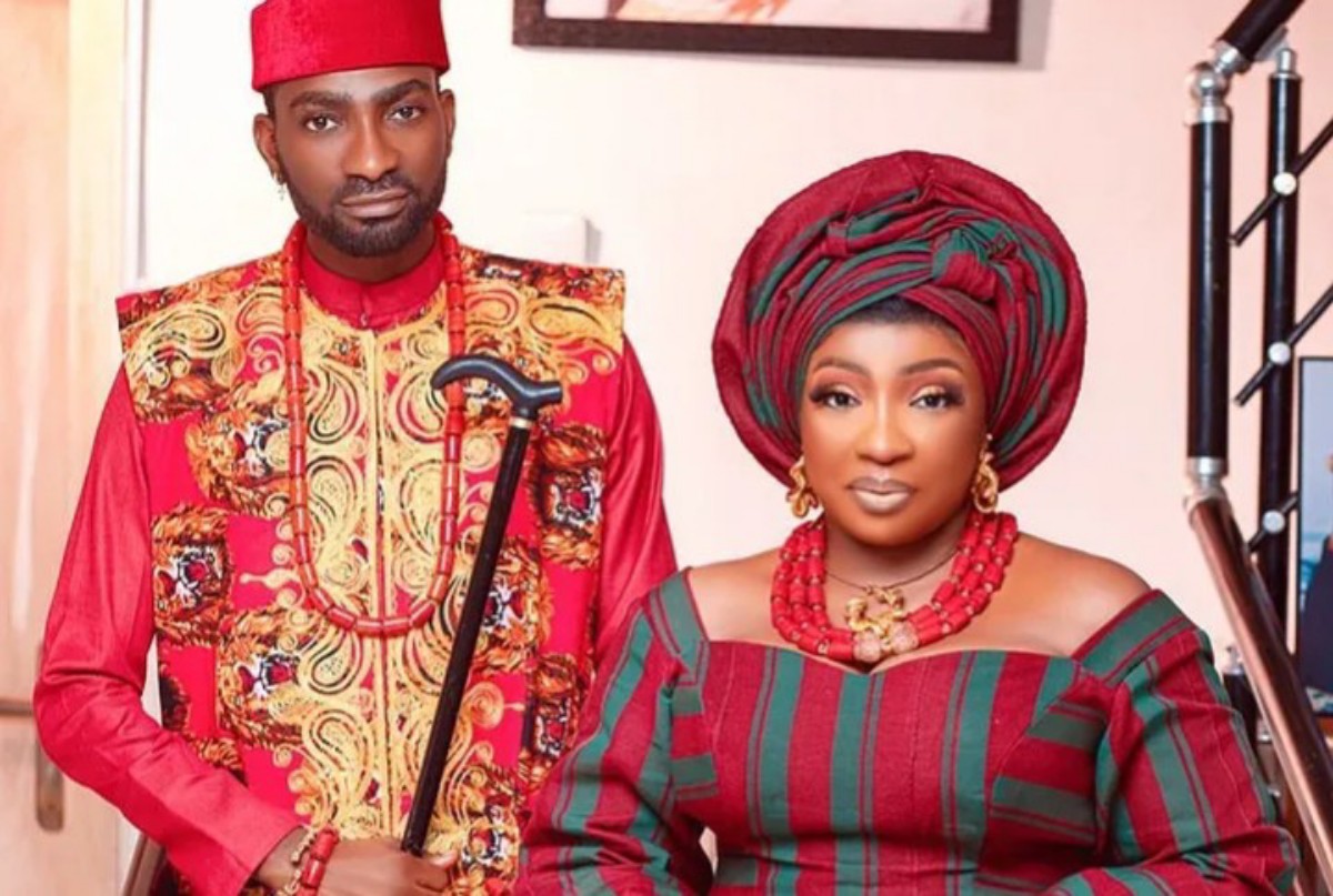 Man and woman in vibrant Nigerian wedding attire posing together; the man holds a decorative staff.