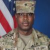 Portrait of a U.S. Army soldier in camouflage uniform with an American flag backdrop in the background.