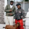 Two smiling people pose outside a building with a brown dog on a leash between them.