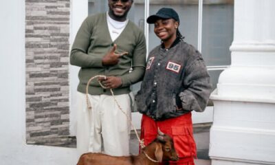 Two smiling people pose outside a building with a brown dog on a leash between them.