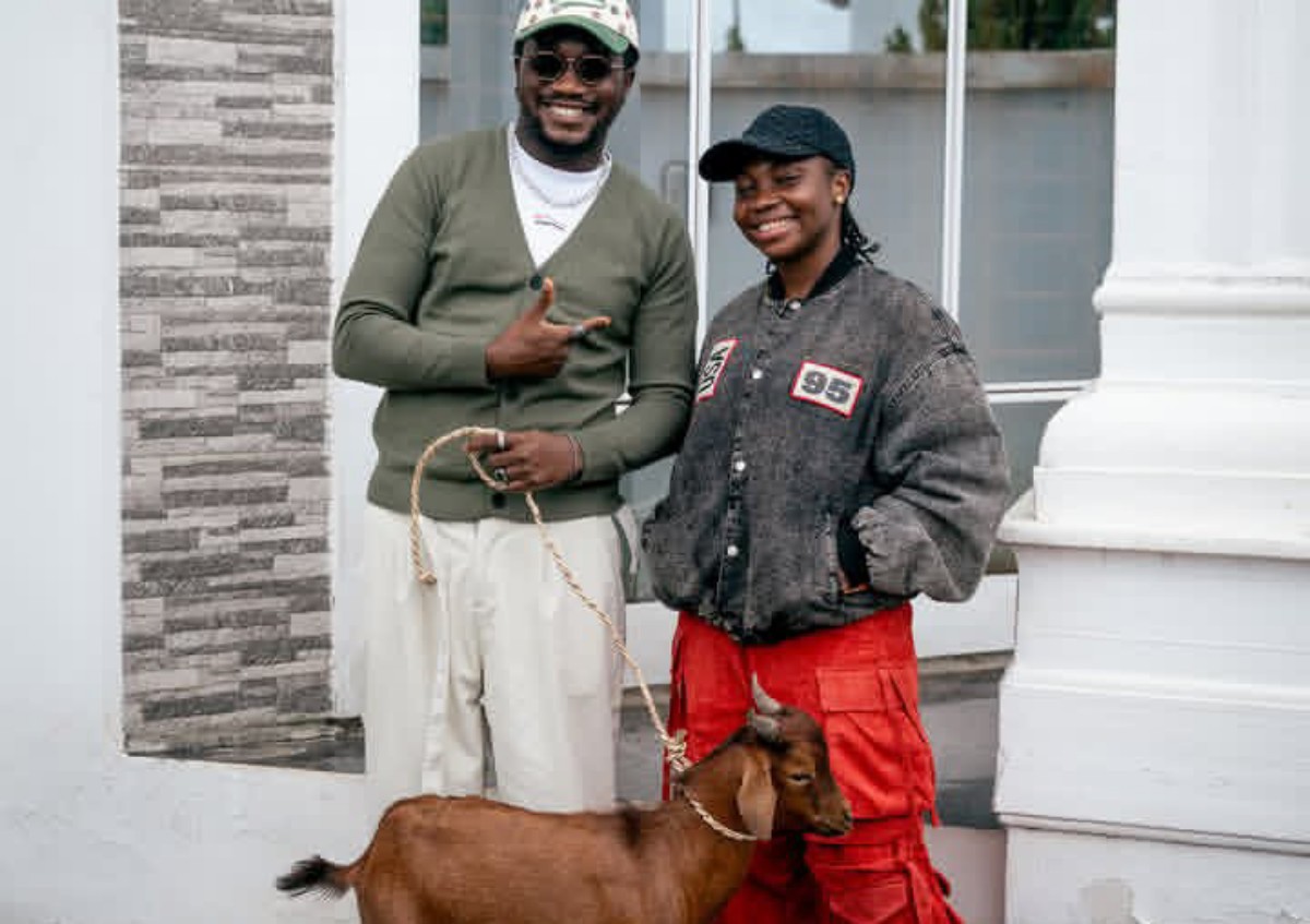 Two smiling people pose outside a building with a brown dog on a leash between them.