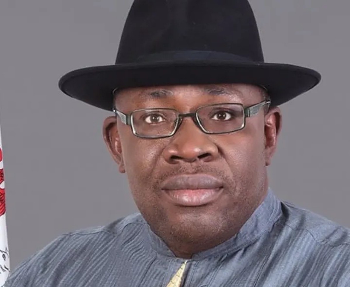 Portrait of a man wearing a black hat, glasses, and a gray tunic, facing the camera against a gray backdrop.