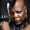 Close-up portrait of a Black person with a mohawk hairstyle, heavy eyeliner, and a nose ring, wearing a studded leather jacket with chains and rings.