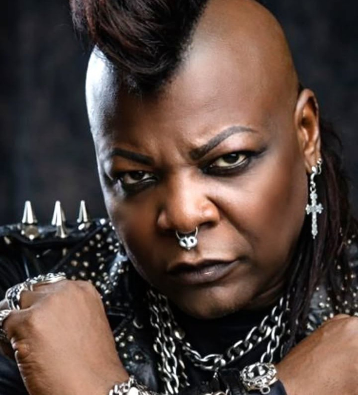 Close-up portrait of a Black person with a mohawk hairstyle, heavy eyeliner, and a nose ring, wearing a studded leather jacket with chains and rings.