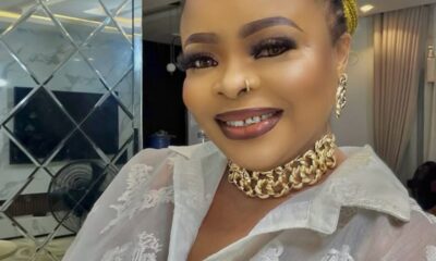 Smiling woman with braided yellow hair, wearing a gold chain choker and sheer white lace blouse indoors.