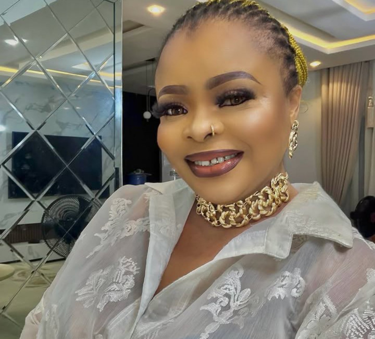 Smiling woman with braided yellow hair, wearing a gold chain choker and sheer white lace blouse indoors.