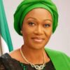Portrait of a woman wearing a bright green headwrap and matching outfit with a gold necklace, posed against a green-and-white background.