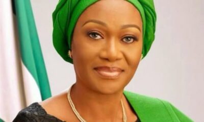 Portrait of a woman wearing a bright green headwrap and matching outfit with a gold necklace, posed against a green-and-white background.