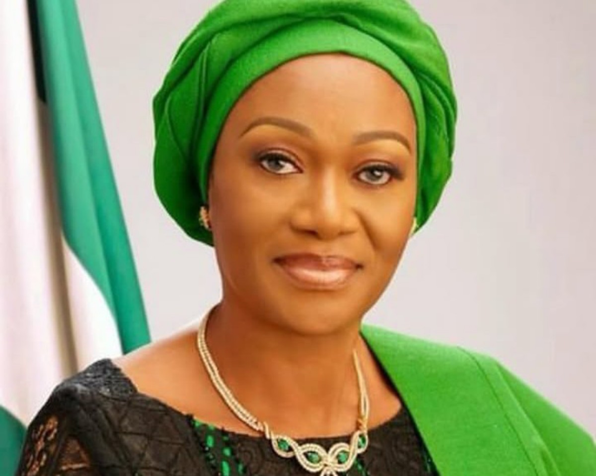 Portrait of a woman wearing a bright green headwrap and matching outfit with a gold necklace, posed against a green-and-white background.