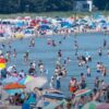 Busy beach filled with people swimming and sunbathing under colorful umbrellas and tents along the shore.