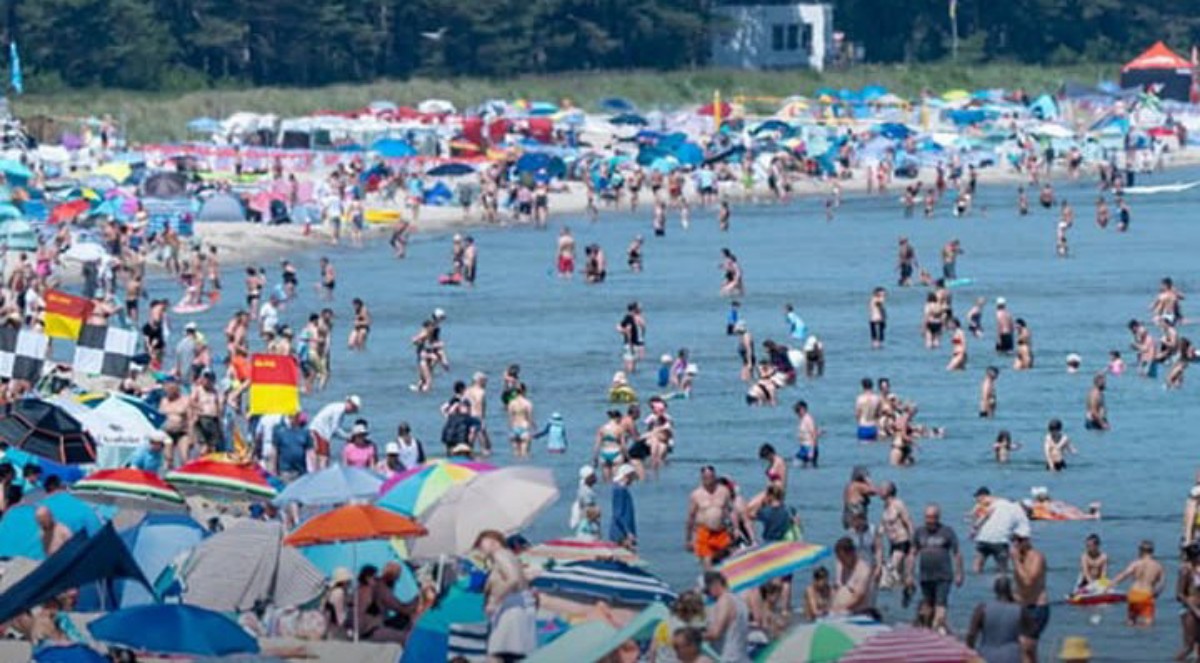 Busy beach filled with people swimming and sunbathing under colorful umbrellas and tents along the shore.