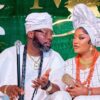 Couple in white traditional Nigerian attire sit together at a wedding, the man holding a staff and wearing a cap while the woman wears a large head wrap and orange bead jewelry with a floral backdrop.