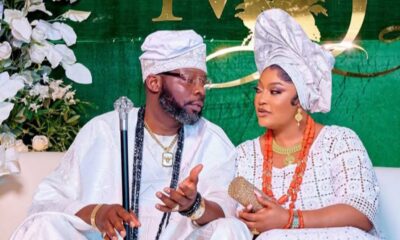 Couple in white traditional Nigerian attire sit together at a wedding, the man holding a staff and wearing a cap while the woman wears a large head wrap and orange bead jewelry with a floral backdrop.
