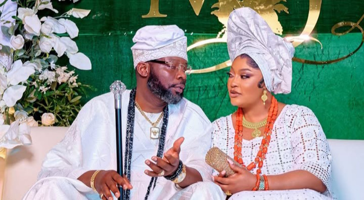 Couple in white traditional Nigerian attire sit together at a wedding, the man holding a staff and wearing a cap while the woman wears a large head wrap and orange bead jewelry with a floral backdrop.