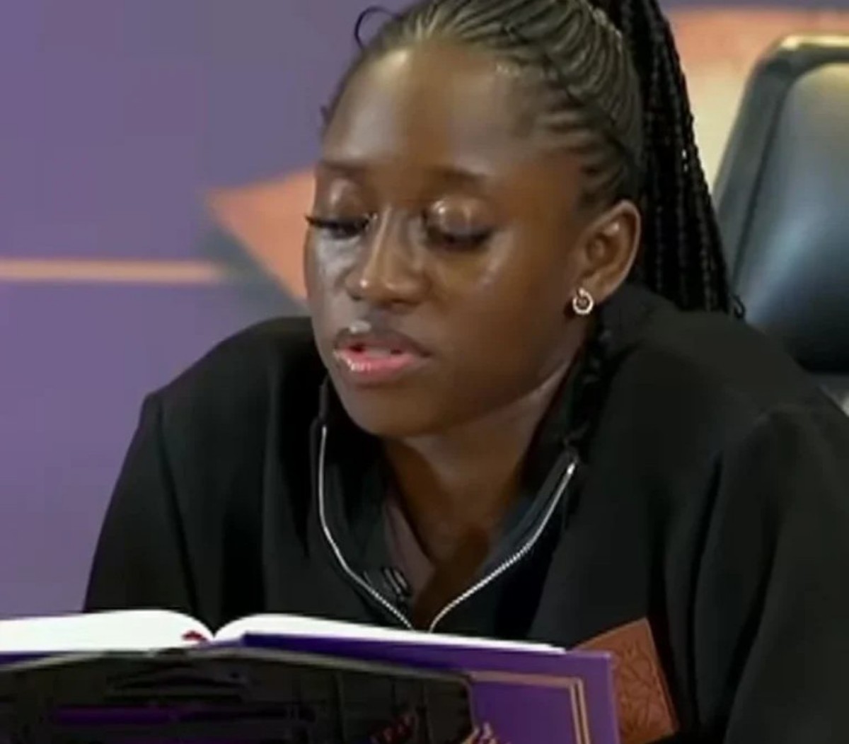Woman with braided hair reading a purple-covered book, wearing a black top, in a studio setting.