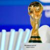 FIFA World Cup trophy close-up on a white pedestal with a blue background, shimmering gold surface visible