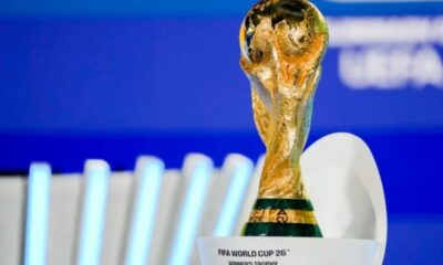 FIFA World Cup trophy close-up on a white pedestal with a blue background, shimmering gold surface visible