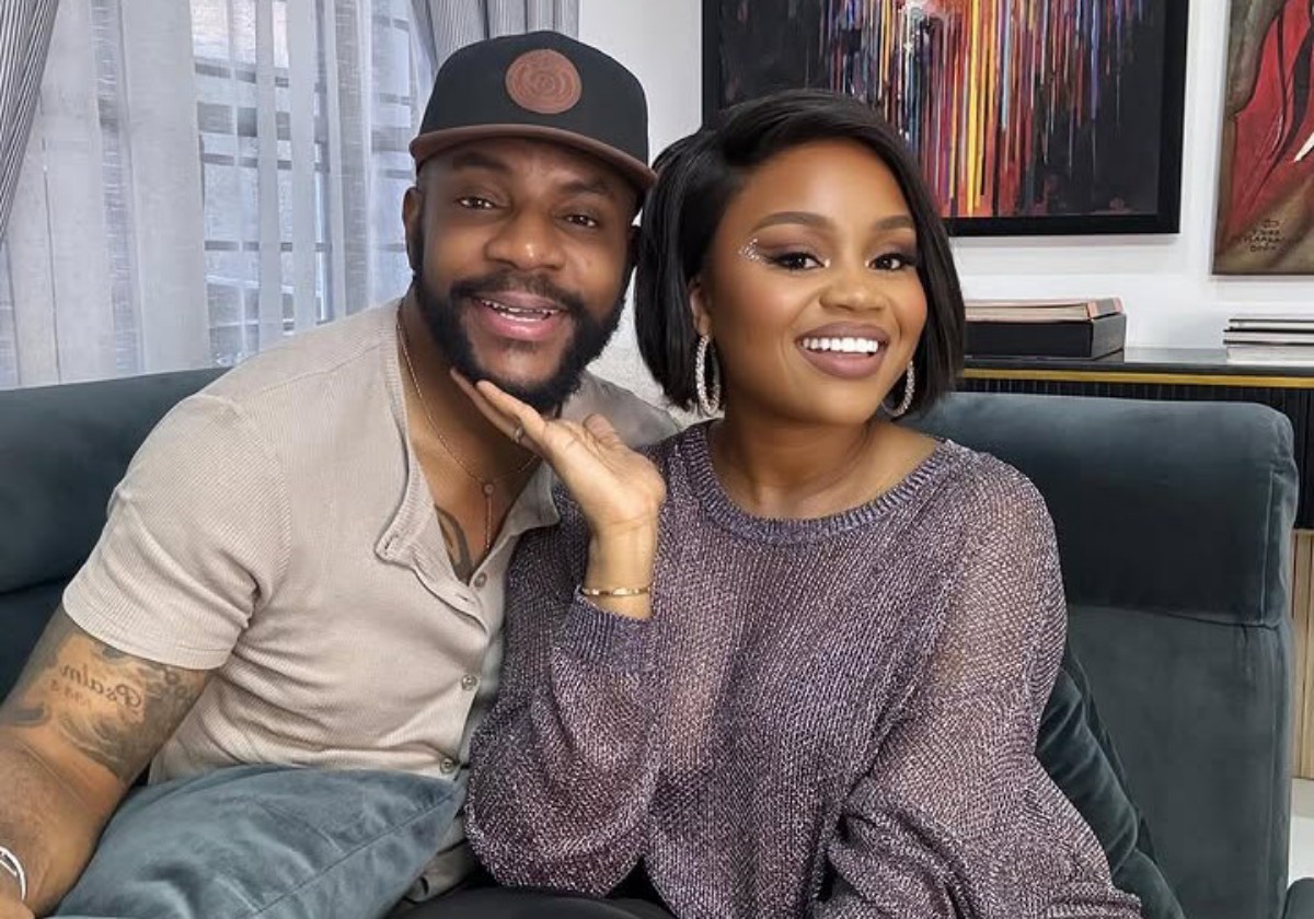A smiling couple sits close together on a dark sofa indoors, posing for a photo with artwork on the wall behind them.