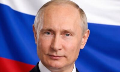 Close-up portrait of a mature man in a dark suit with a white shirt, Russia's flag in the background.