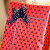 Red gift box with black polka dots, tied with a dark bow on top and a light gray ribbon crossing the front.