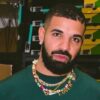 Close-up portrait of a man with a trimmed beard wearing a green shirt and a colorful beaded necklace that says 'LOVE', standing in front of stacked sneaker boxes in a warehouse setting.
