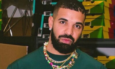Close-up portrait of a man with a trimmed beard wearing a green shirt and a colorful beaded necklace that says 'LOVE', standing in front of stacked sneaker boxes in a warehouse setting.