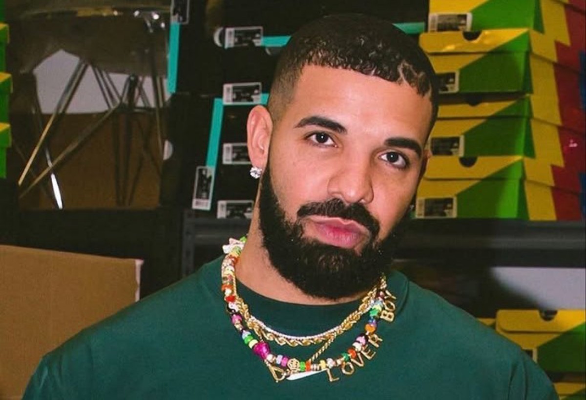 Close-up portrait of a man with a trimmed beard wearing a green shirt and a colorful beaded necklace that says 'LOVE', standing in front of stacked sneaker boxes in a warehouse setting.