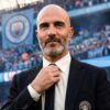 Man in a black suit adjusts his white tie in a stadium packed with blue-clad fans, Manchester City crest on his blazer visible?