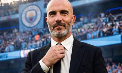 Man in a black suit adjusts his white tie in a stadium packed with blue-clad fans, Manchester City crest on his blazer visible?