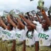 Line of Nigerian women in NYSC uniforms raising hats in a salute during a parade would be the best substitute for ALT text here, capturing the scene and purpose without unnecessary visuals.