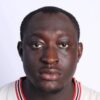 Close-up portrait of a man with a neutral expression, wearing a white jersey with red/black trim against a white background