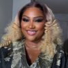 Smiling person with blonde, voluminous curly hair wearing a metallic floral robe and a gold necklace.