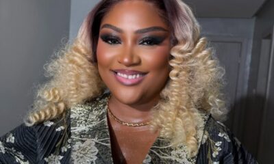 Smiling person with blonde, voluminous curly hair wearing a metallic floral robe and a gold necklace.