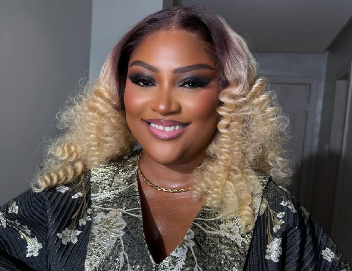 Smiling person with blonde, voluminous curly hair wearing a metallic floral robe and a gold necklace.