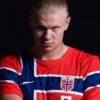 A young man with slicked-back blond hair wearing a red, white, and blue Norway football jersey with crest, posed against a dark background.