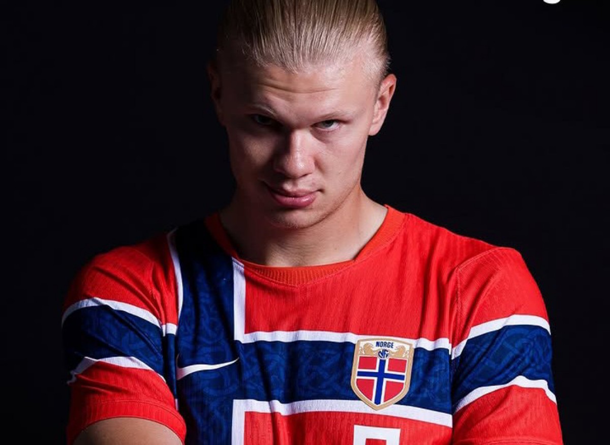 A young man with slicked-back blond hair wearing a red, white, and blue Norway football jersey with crest, posed against a dark background.