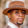 Portrait of a man wearing a brown felt hat and round sunglasses, looking over his shoulder.