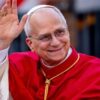 Pope Francis smiles and waves, dressed in bright red papal robes and a white skullcap.