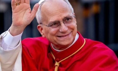 Pope Francis smiles and waves, dressed in bright red papal robes and a white skullcap.