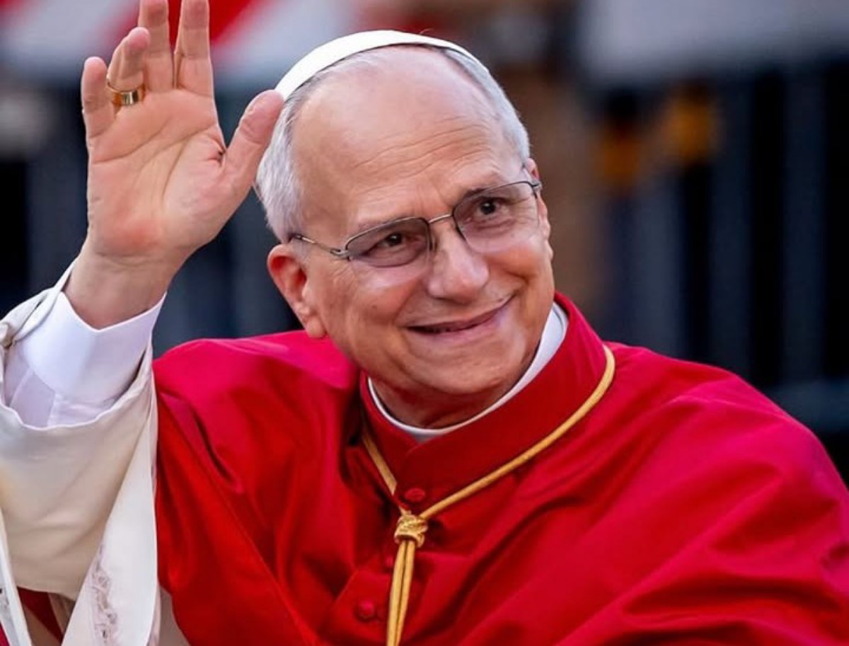 Pope Francis smiles and waves, dressed in bright red papal robes and a white skullcap.