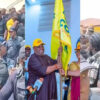 A crowd of people at a political event, with a central figure holding a yellow flag and smiling for cameras.