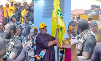 A crowd of people at a political event, with a central figure holding a yellow flag and smiling for cameras.