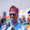 Man wearing a red-and-navy traditional cap and sunglasses speaks into multiple microphones at a press conference, with a uniformed officer in a blue beret behind him.
